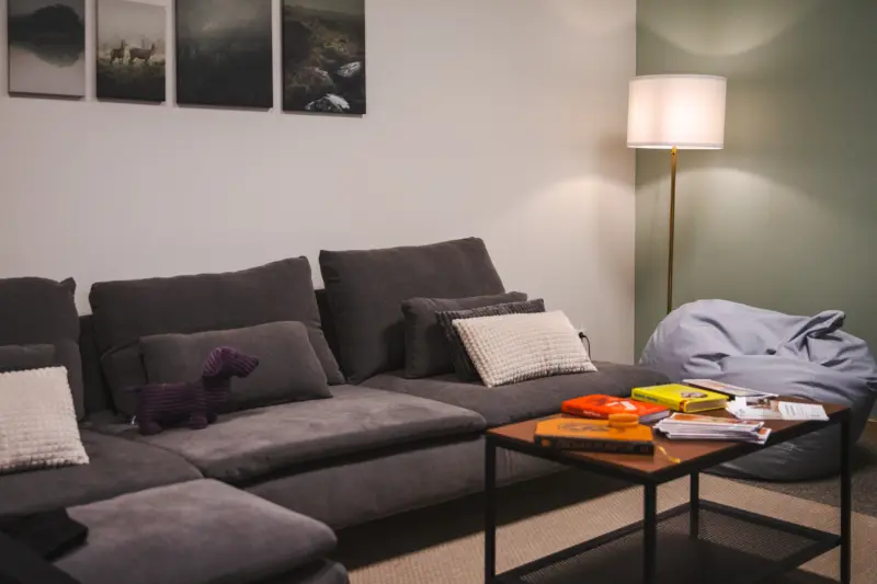 Ett rum möblerat med en svart soffa fylld med kuddar och ett gosedjur, en tavelvägg ovanför soffan, en golvlampa, ett soffbord och en grå saccosäck till höger om soffan.