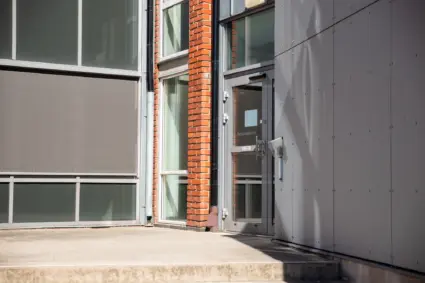 En exteriörbild på en entrédörr i glas. Fasaden till byggnaden är i grå plåt samt rött tegel.