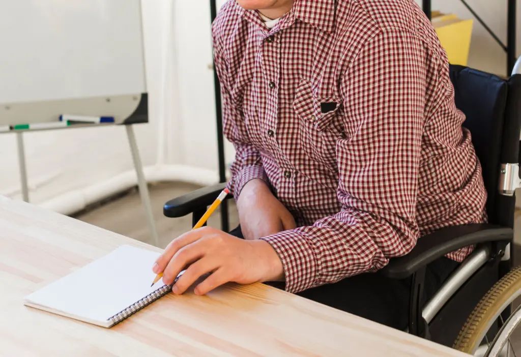 Man i rullstol sitter vid bord och skriver i ett anteckningsblock
