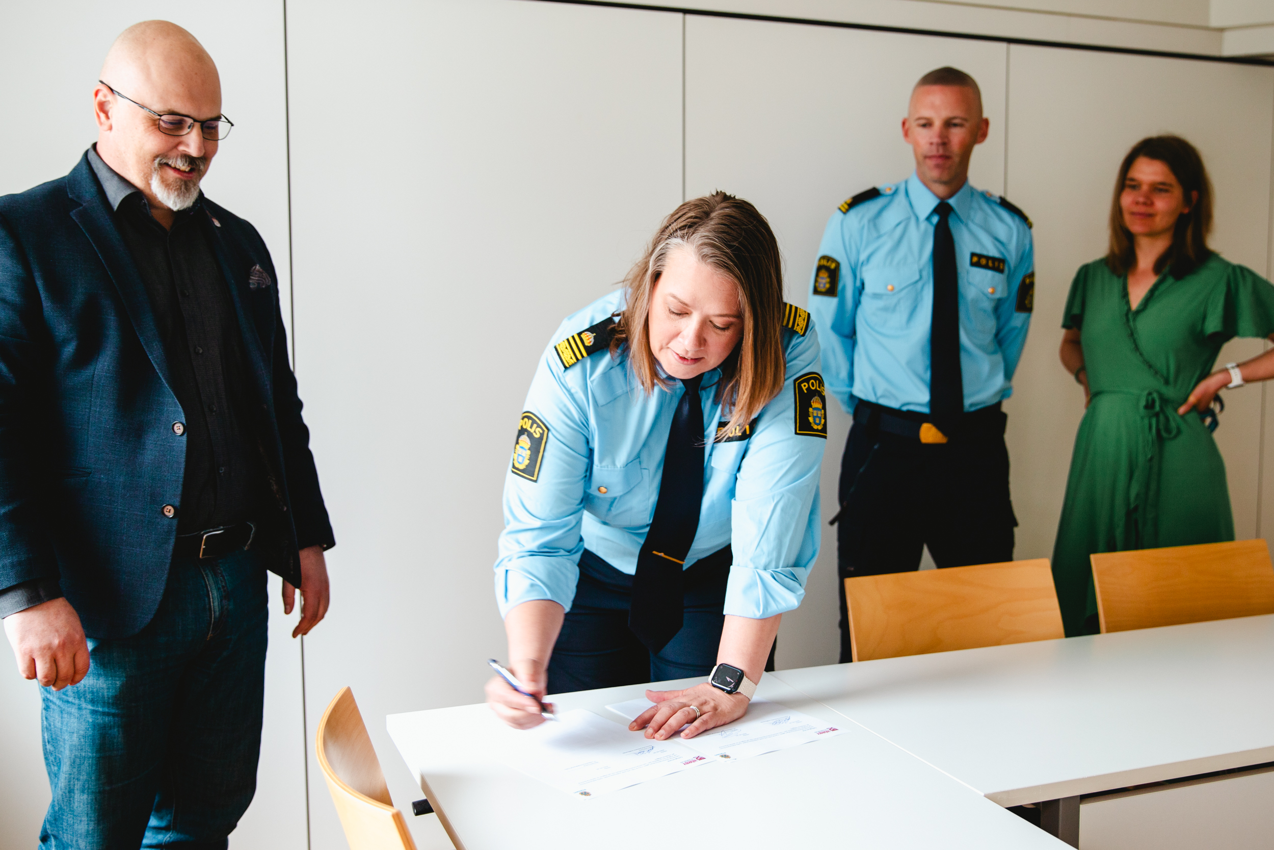 Fyra personer syns i bilden, som är tagen i ett ljust rum inomhus. En av personerna, en kvinna, är i full gång att skriva under ett papper. De andra bevittnar henne samtidigt som de ler.