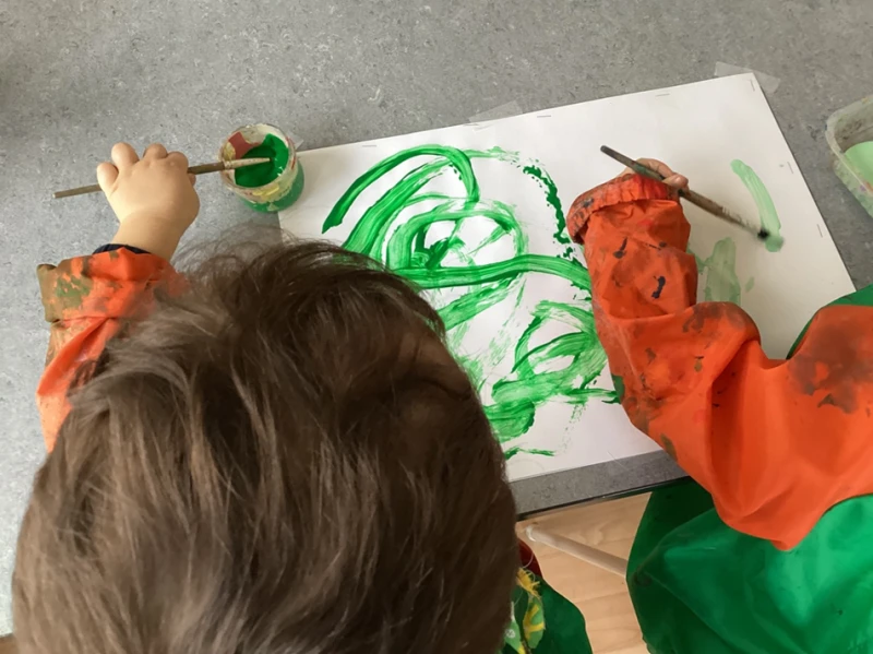 Bild ovanifr&aring;n p&aring; tv&aring; barn som m&aring;lar med gr&ouml;n m&aring;larf&auml;rg p&aring; en vit canvas,