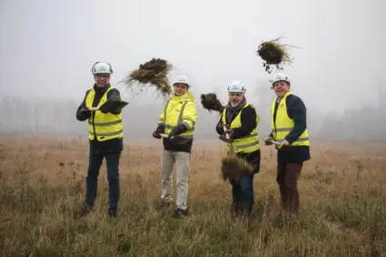 Fyra män klädda i gul varselväst står på en dimmig åker en mulen dag. De håller i varsin spade som de har använt för att ta ett symboliskt första spadtag.
