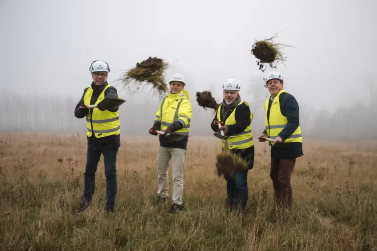 Fyra män klädda i gul varselväst står på en dimmig åker en mulen dag. De håller i varsin spade som de har använt för att ta ett symboliskt första spadtag.