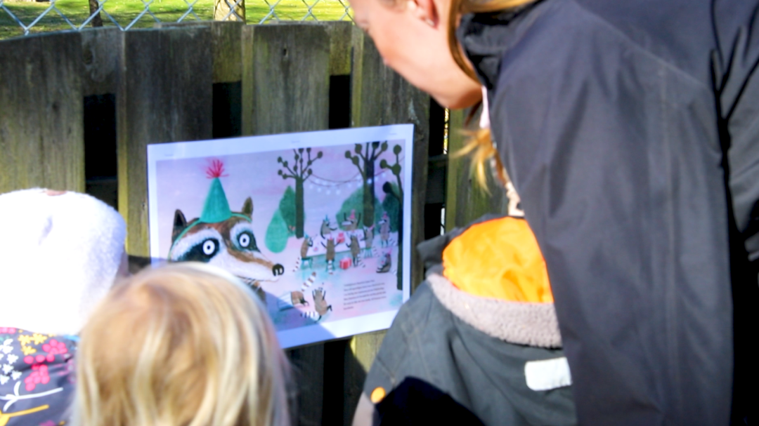 Tre barn och en pedagog står och tittar på en illustration som hänger på ett staket.