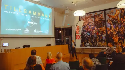 En kvinna står framför ett podie och pratar i en mikrofon för en grupp med människor som sitter på stolar.
