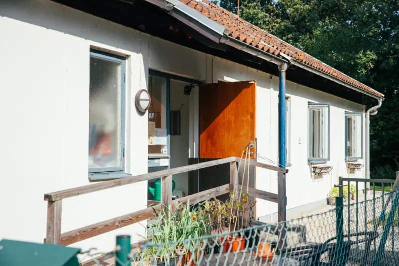 Halvbild på exteriören av ett vitt hus. I mitten av bilden syns en öppen trädörr, vilken en ramp leder upp till. Framför rampen syns tomatplantor och andra växter i krukor. Det är en solig dag