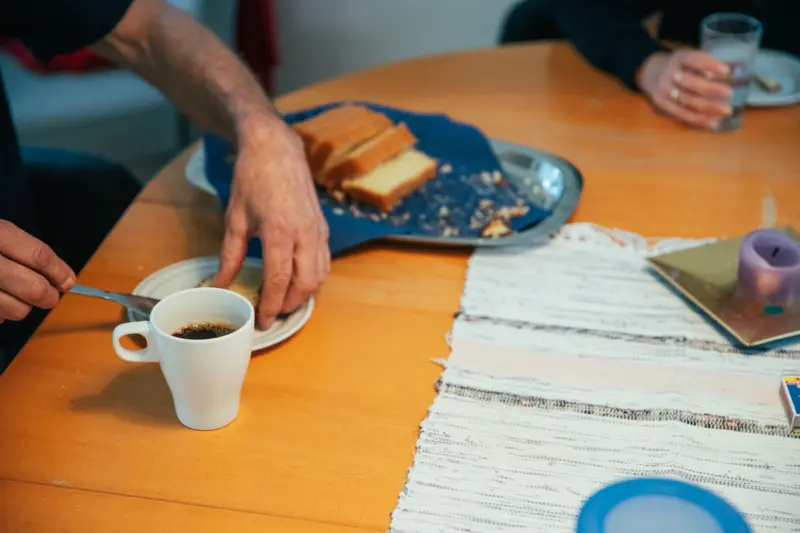En halvbild på ett köksbord. På bordet står en kaffekopp med svart kaffe, samt ett silverfat med uppskivad sockerkaka. Ett par händer håller på att lägga upp en skiva kaka på en mindre assiett.