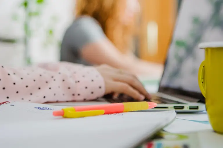 Närbild på flickas arm som skriver på en laptop, med penna och anteckningsblock i förgrunden